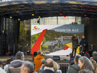 Bühne mit großem Bildschirm und Aufschrift „Reisen für Alle“, Moderator spricht mit einem Mann im Rollstuhl, Publikum davor.