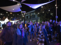 Menschenmenge auf einem Platz bei Nacht unter Lichterketten, Marktstände im Hintergrund.