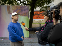 Kamerateam führt Interview mit einem Mann in blauem Pullover, Frau hält Mikrofon mit ZDF-Logo.