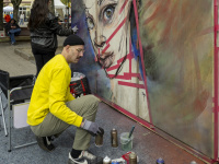 Künstler arbeitet mit Spraydosen an einem großflächigen Wandbild mit Frauengesicht, weitere Person im Hintergrund.