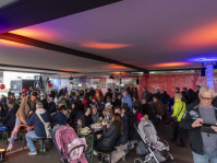 Überdachter Veranstaltungsbereich mit langen Tischen und vielen Besuchern.