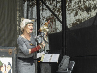 Frau mit Kopfbedeckung zeigt Handpuppen auf einer Bühne vor einem schwarzen Hintergrund.