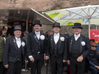 Vier Männer in schwarzem Frack mit Zylinder und weißen Blumen posieren gemeinsam vor einem Stand.