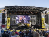 Auf einer Bühne stehen drei Leute, vor der Bühne viele Zuschauer.