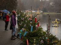 Menschen laufen an einer Reihe kleiner geschmückter Tannenbäume am Seeufer entlang; im Wasser leuchtet ein großer Stern, im Hintergrund sind weitere Marktstände zu sehen.