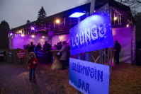Eine Glühwein-Lounge mit lila Beleuchtung, in der mehrere Personen an Stehtischen stehen; vorne ein Kind, das Pommes isst, auf einem mit Stroh bedeckten Boden.