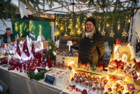 Eine Händlerin steht hinter einem weihnachtlichen Keramikstand mit Miniaturhäusern, roten Dekofiguren und vielen warm leuchtenden Lichtern.