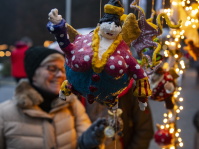 Eine bunte, handgefertigte, hängende Figur in Nahaufnahme, dahinter unscharf eine Frau, die den Marktstand betrachtet, umgeben von warmen Lichterketten.