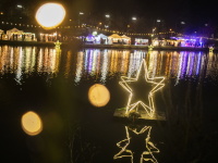Leuchtende Sterninstallation auf dem Wasser in der Nacht, im Hintergrund bunte Marktstände und Lichterreihen, deren Reflexe sich auf der Wasseroberfläche spiegeln.