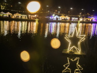 Leuchtende Sterninstallation auf dem Wasser in der Nacht, im Hintergrund bunte Marktstände und Lichterreihen, deren Reflexe sich auf der Wasseroberfläche spiegeln.