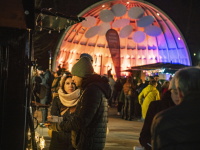 Ein Paar steht vor einer bunt beleuchteten Bühne, umgeben von Besuchern im warmen Winterlicht.