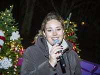 Eine lächelnde Frau spricht ins Mikrofon, umgeben von weihnachtlich geschmückten Tannen.