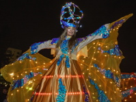 Eine Stelzenläuferin trägt ein leuchtendes, farbenprächtiges Kostüm im Dunkeln.