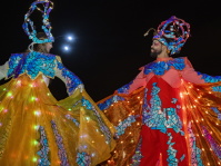 Zwei Stelzenläufer in bunten, leuchtenden Kostümen tanzen einander gegenüber.