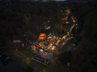 Luftaufnahme zeigt Bühne, Buden und Wege eines hell beleuchteten Weihnachtsmarkts im Wald.
