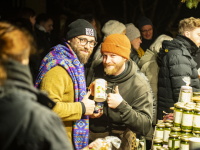Zwei Männer lachen in die Kamera und zeigen Getränke, dicht gedrängt zwischen Marktbesuchern.