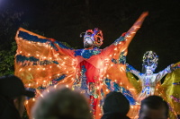 Zwei Künstler auf Stelzen tragen leuchtende, farbenfrohe Kostüme und breiten ihre illuminierten Flügel vor dem Publikum aus.