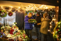 ine Gruppe betrachtet Adventsdekoration an einem Stand, während Lichterketten den Platz erhellen.