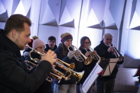 Eine Bläsergruppe spielt gemeinsam festliche Musik, dicht aneinandergereiht unter kühlem Bühnenlicht.