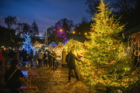 Besucher schlendern zwischen festlich beleuchteten Holzbuden und bunt geschmückten Tannenbäumen über den Weihnachtsmarkt in der Dämmerung.