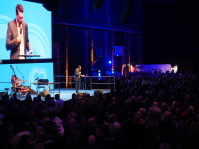 Mann auf Bühne mit großer Leinwand, die ihn zeigt