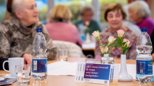 Im Hintergrund mehrere Senioren in einer Veranstaltung, im Vordergrund ein Tisch mit Flyern zur Kampagne 
