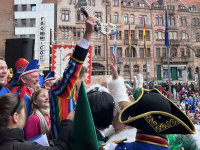 Mehrere kostümierte Personen stehen auf einer Bühne, während ein Mann im bunten Kostüm einen großen Schlüssel in die Luft hält.