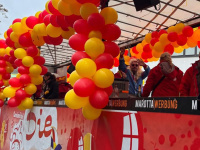 Ein Umzugswagen ist mit roten und gelben Luftballons dekoriert, während mehrere Personen auf dem Wagen stehen und feiern.