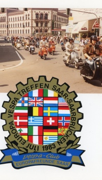 Oben ist ein Foto mit zahlreichen Vespa-Fahrern zu sehen, die an der Bergwerksdirektion in Saarbrücken vorbeikommen. Unten ist ein Sticker des Vespa-Clus Eurpafreunde Saar anlässlich des Europa Vespa-Treffens In Saarbrücken im Juli 1983 zu sehen.