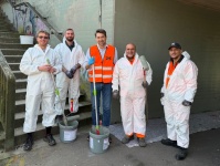 Fünf Personen in weißen Schutzanzügen und orangen Warnwesten stehen mit Farbrollern und Farbeimern vor einer  Unterführung