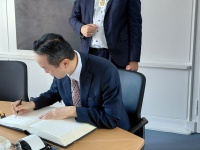 Mann schreibt an einem Tisch in ein Gästebuch, dahinter steht ein weiterer Mann mit Amtskette vor einem großformatigen Gemälde.