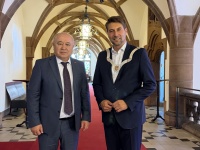 Zwei Männer in Anzügen stehen auf einem roten Teppich in einem historischen Flur mit Gewölbedecke und großen Hängeleuchten.