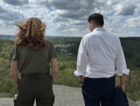 Eine Frau und ein Mann stehen mit dem Rücken zur Kamera auf einer Anhöhe und blicken über eine bewaldete Landschaft.