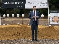 Ein Mann im Anzug steht auf einer Baustelle vor einer Reihe von Spaten mit weißen Bauhelmen und hält eine Ansprache.