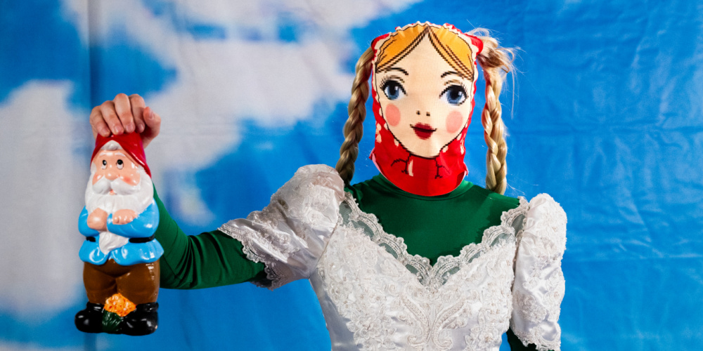 Eine Person in einem Brautkleid trägt eine Matroschka-Maske und hält einen Gartenzwerg in der Hand 