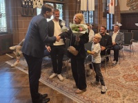 Ein Mann schüttelt einer Frau mit Blumenstrauß die Hand. Mehrere Personen sitzen im Hintergrund eines historischen Saals.