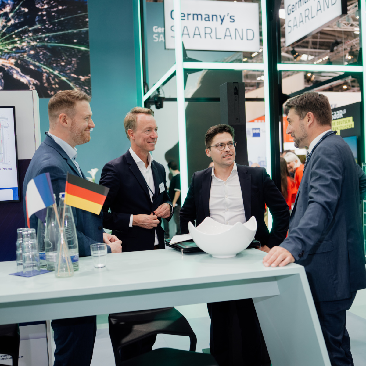 Vier Männer stehen an einem Hochtisch am Messestand und unterhalten sich. Auf dem Tisch steht eine Vase mit einem deutschen und einem französischen Fähnchen.